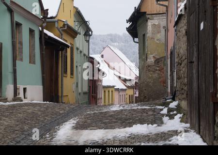 Besucher spazieren entlang der kopfsteingepflasterten Straßen der Zitadelle Sighisoara, umgeben von farbenfrohen Gebäuden und einer leichten Schneeschicht. Stockfoto