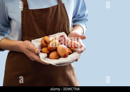 Eine Person, die eine braune Schürze trägt, hält einen Teller mit dekorierten Ostereiern. Die Eier zeigen rustikale Muster und Farben, die eine handgefertigte Ästhetik verkörpern. Th Stockfoto