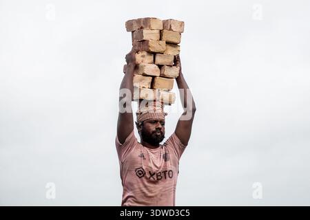 Satkhira, Bangladesch - 25. April 2025: Blick auf einen Mann, der einen Stapel Ziegeln auf seinem Kopf trägt, seine Figur eine Silhouette vor dem hellen Himmel. Stockfoto