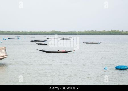 Satkhira, Bangladesch - 25. April 2025: Blick auf traditionelle Holzboote, die sanft auf dem ruhigen, grauen Wasser schwimmen und den bewölkten Himmel in der Nähe des reflektieren Stockfoto