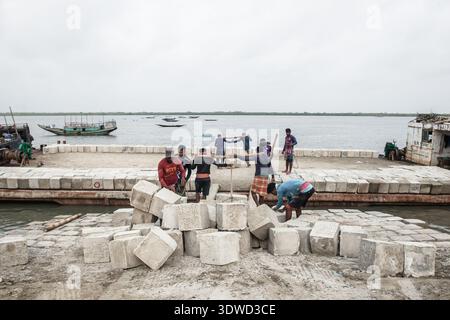 Satkhira, Bangladesch - 25. April 2025: Blick auf Arbeiter, die einen stabilen, grauen Damm entlang der ruhigen, weitläufigen Küste bauen, mit Fischen bo Stockfoto