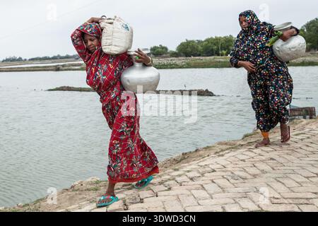 Satkhira, Bangladesch - 25. April 2025: Blick auf zwei Frauen in leuchtenden Blumenkleidern mit Wasserkannen, deren Reflexionen auf der ruhigen Wa glitzern Stockfoto