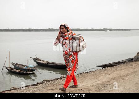 Satkhira, Bangladesch - 25. April 2025: Blick auf eine Frau in lebendiger Blumenbekleidung mit einem metallischen Topf am Ufer, in der Nähe des traditionellen hölzernen bo Stockfoto