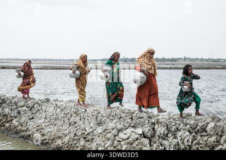 Satkhira, Bangladesch - 25. April 2025: Blick auf Frauen, die einen schmalen, felsigen Pfad inmitten der überfluteten Landschaft durchqueren, darunter jeweils ein Metallschiff Stockfoto