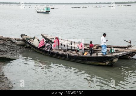 Satkhira, Bangladesch - 25. April 2025: Blick auf hölzerne Boote, die Menschen entlang der schlammigen Flussufer transportieren und einen düsteren grauen Himmel und das tägliche Leben widerspiegeln Stockfoto