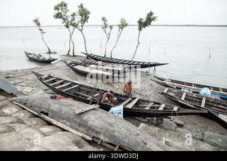Satkhira, Bangladesch - 25. April 2025: Blick auf hölzerne Boote, die auf dem matschigen Ufer ruhen, deren dunkle Rümpfe sich gegen den hellen Himmel und noch Wasser abheben Stockfoto