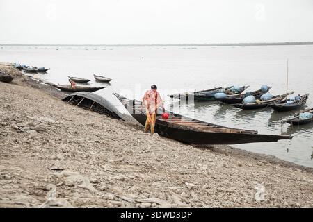 Satkhira, Bangladesch - 25. April 2025: Blick auf eine Frau in einem leuchtenden orange-roten Sari, die an einem dunklen Holzboot am schlammigen Ufer des Flusses steht. Stockfoto