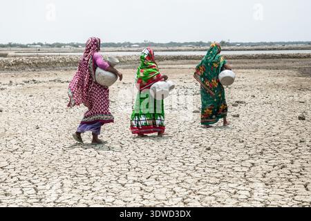 Satkhira, Bangladesch - 25. April 2025: Blick auf Frauen, die die trockene, zerrissene Erde einer Salzpfanne überqueren und unter dem hellen Himmel schwere Belastungen tragen. Stockfoto