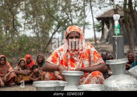 Satkhira, Bangladesch - 25. April 2025: Blick auf eine Frau in einem orange-weißen blumigen Hijab, der zwischen silbernen Töpfen sitzt, die Wasserpumpe liefert sauberes Wasser Stockfoto