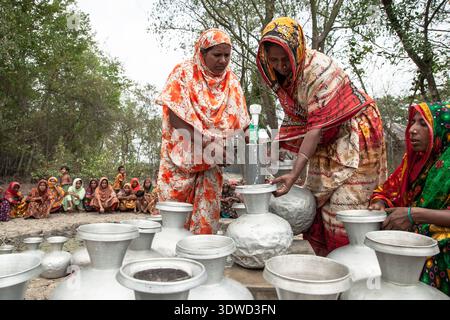 [Satkhira], [Bangladesch] - 25. April 2025: Blick auf Frauen, die Wasser in Aluminiumtöpfen unter freiem Himmel sammeln, umgeben von üppigem Grün und Erdboden Stockfoto