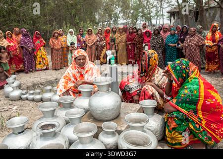 [Satkhira], [Bangladesch] - 25. April 2025: Blick auf Frauen, die sich um glänzende Metalltöpfe versammeln. Ihre lebendigen Saris bilden einen krassen Kontrast zu dem trockenen, staubigen Gräuer Stockfoto