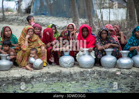 Satkhira, Bangladesch - 25. April 2025: Blick auf Frauen und Kinder, die mit Metallgefäßen an einer schlammigen Bank sitzen, inmitten des sanften Lichts und der Erdtöne. Stockfoto