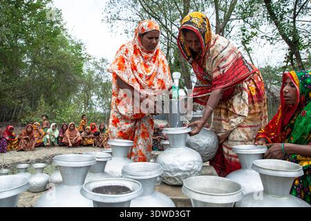 Satkhira, Bangladesch - 25. April 2025: Blick auf Frauen in farbenfrohen Saris, die Metallkrug mit Wasser aus einer Pumpe füllen und das Licht gegen eine Ba reflektieren Stockfoto
