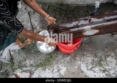 Satkhira, Bangladesch - 14. Juli 2025: Blick auf die Hände einer Frau mit einem rostigen Metallblech, um Wasser aus einem silbernen Topf in einen roten Eimer zu gießen Stockfoto