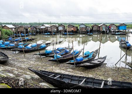 Satkhira, Bangladesch - 13. Juli 2025: Blick auf traditionelle Holzboote auf dem schlammigen Ufer, die die rustikalen Pfahlhäuser am Wasser reflektieren Stockfoto
