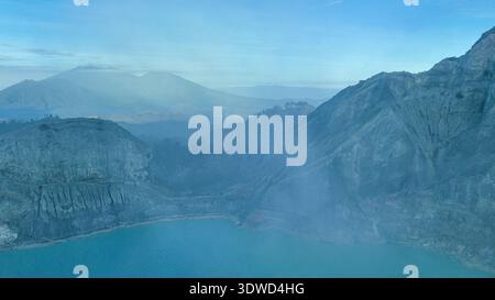 Türkisfarbenes Wasser des Kawah Ijen Acid Lake wirbelt unter zerklüfteten Kraterwänden und schwimmenden Schwefelwolken und erzeugt einen eindringlich schönen vulkanischen Amphit Stockfoto
