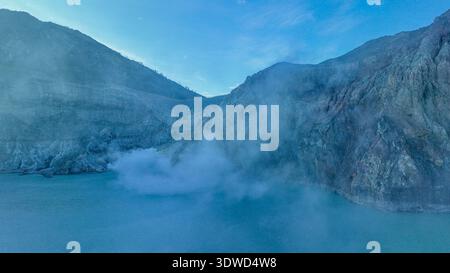 Türkisfarbenes Wasser des Kawah Ijen Acid Lake wirbelt unter zerklüfteten Kraterwänden und schwimmenden Schwefelwolken und erzeugt einen eindringlich schönen vulkanischen Amphit Stockfoto