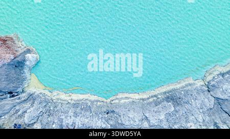 Türkisfarbenes Wasser des Kawah Ijen Acid Lake wirbelt unter zerklüfteten Kraterwänden und schwimmenden Schwefelwolken und erzeugt einen eindringlich schönen vulkanischen Amphit Stockfoto