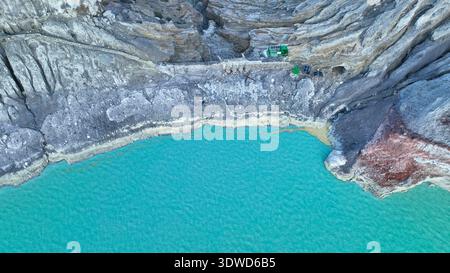 Türkisfarbenes Wasser des Kawah Ijen Acid Lake wirbelt unter zerklüfteten Kraterwänden und schwimmenden Schwefelwolken und erzeugt einen eindringlich schönen vulkanischen Amphit Stockfoto