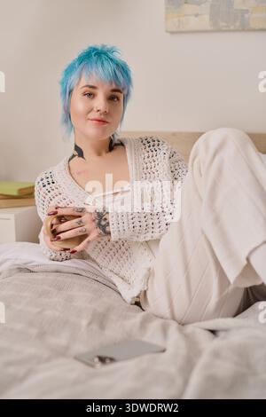 Die lebendige Frau in einem Strickoberteil entspannt sich mit einem Becher, während sie auf ihrem Bett liegt. Stockfoto
