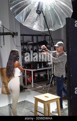 Ein Model posiert für ein Fotoshooting in einem Studio, während ein Fotograf sie mit einer Kamera festnimmt. Der Raum ist mit professioneller Beleuchtung und Requisiten ausgestattet Stockfoto
