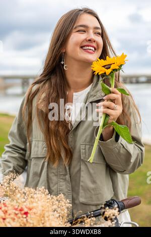 Eine Frau in einem Park lächelt fröhlich, während sie eine helle Sonnenblume in der Nähe ihres Gesichts hält, mit einem Fahrrad und trockenen Blumen im Vordergrund. Stockfoto