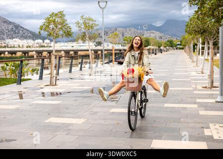 Eine fröhliche Frau fährt mit dem Fahrrad einen Parkweg entlang, ihr Korb ist voller bunter Blumen. Ihr Ausdruck strahlt Freude aus, während sie das schöne Ou genießt Stockfoto