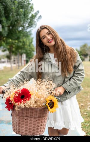 Eine fröhliche Frau mit langen Haaren lächelt, während sie durch einen üppigen Park radelt, ihr Fahrradkorb voller lebendiger Blumen. Sie trägt eine leichte Jacke und eine Stockfoto