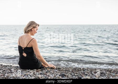 Eine ruhige Szene einer Frau in schwarzem Kleid, die an einem Kiesstrand sitzt, während sie während des Sonnenuntergangs auf die beruhigenden Wellen des Ozeans blickt und einen Moment der Ruhe einnimmt Stockfoto