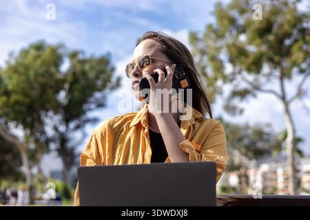 Frau im Park, die einen Laptop benutzt und telefoniert, verkörpert den Lebensstil der Fernarbeit. Lässig gekleidet, bringt sie Technologie und Natur nahtlos in Einklang Stockfoto