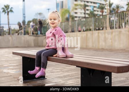 Ein fröhliches Kleinkind sitzt auf einer Holzbank, trägt einen rosa Pullover und Socken neben ihren rosa Gummistiefeln und genießt einen regnerischen Tag im Freien in einem urb Stockfoto