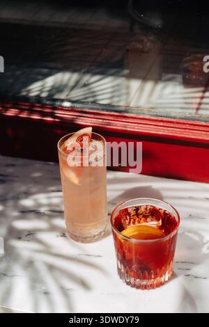 Ein erfrischender Negroni und ein Grapefruit-Cocktail werden elegant auf einem Marmortisch platziert. Sonnenlicht wirft Schatten und hebt die ästhetische Szene hervor Stockfoto