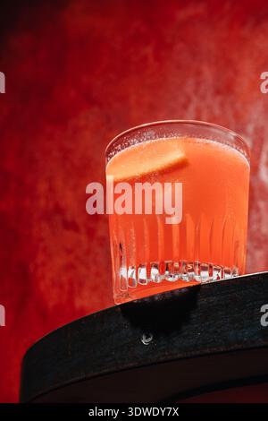Ein erfrischender, oranger Negroni in einem strukturierten Glas, vor einem auffälligen roten Hintergrund. Das lebhafte Getränk wird mit einer Scheibe Orangenruhe garniert Stockfoto