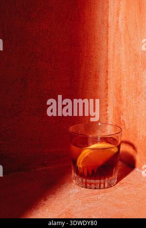 Ein klassischer Negroni-Cocktail in einem Glas, garniert mit Orangenscheibe, auf einer strukturierten roten Oberfläche. Die tiefen Töne verstärken die reichhaltige Ästhetik Stockfoto