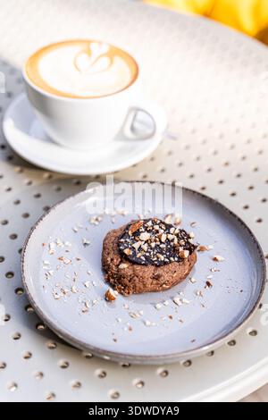 Ein köstlicher Cappuccino sitzt in einer weißen Tasse, begleitet von einem Schokoladenkeks mit Nüssen auf einem grauen Teller, der auf einem perforierten Kaffeetisch steht Stockfoto