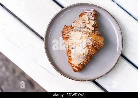 Ein frisches Mandelcroissant sitzt auf einer grauen Keramikplatte, elegant auf einem rustikalen weißen Holztisch angeordnet. Seine Oberfläche ist mit Puderzucker an bestäubt Stockfoto