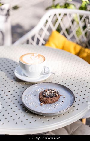 Eine herrliche Café-Szene im Freien mit einem schaumigen Cappuccino in einer weißen Tasse und einem Schokoladenkeks mit Nüssen, auf einem runden Tisch mit einem W Stockfoto