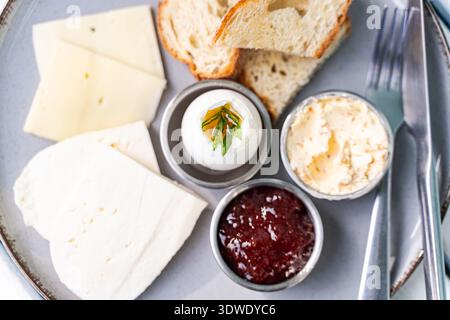 Ein köstlicher Frühstücksteller mit frischen Brotscheiben, zwei Käsesorten, einem gekochten Ei garniert mit Kräutern, cremiger Butter und einer süßen Marmelade für Stockfoto