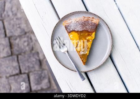 Ein köstliches Stück Mangobutte auf einem runden Teller, auf einem weißen Tisch. Der Kuchen hat eine goldene Kruste und eine lebendige Mangofüllung Stockfoto
