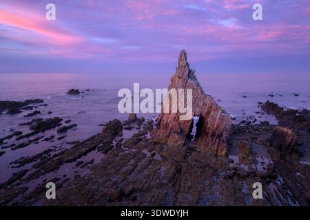 Ein atemberaubender Blick auf eine prächtige Felsformation, die in das ruhige Meer ragt, die Silhouette vor einem leuchtenden rosa und violetten Sonnenuntergangshimmel, Showcasin Stockfoto