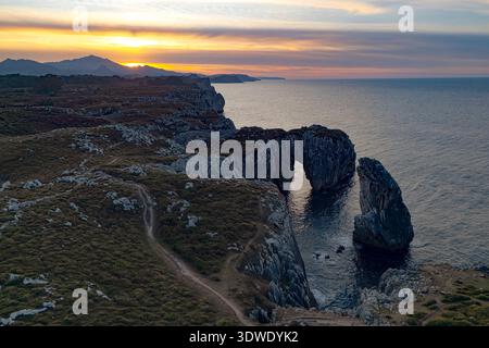 Eine atemberaubende Küstenlandschaft bei Sonnenuntergang mit zerklüfteten Klippen und einem natürlichen Felsbogen, der sich bis ins Meer erstreckt. Die heitere Szene ist in warm getaucht, Stockfoto