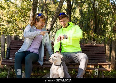 Geschwister genießen einen friedlichen Moment auf einer Parkbank mit ihrem Hund. Sie lachen, während sie ihre Smartwatches überprüfen, um die Freude der Natur und zu genießen Stockfoto