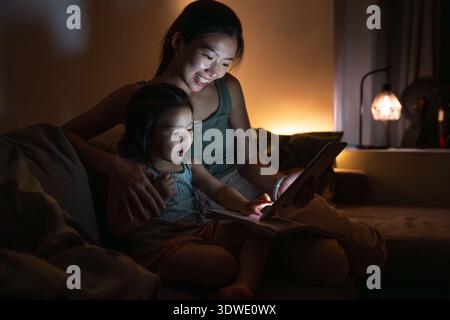 Mutter und Tochter teilen sich einen gemütlichen Moment auf dem Sofa, beleuchtet vom warmen Leuchten einer Lampe und dem Bildschirm eines digitalen Tablets. Das Zimmer ist dunkel Stockfoto