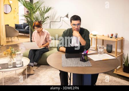 Zwei junge Berufstätige arbeiten in einem modernen Coworking Office. Der eine benutzt einen Laptop, der andere macht eine Pause mit einem Drink. Der Raum fördert die Kreativität Stockfoto