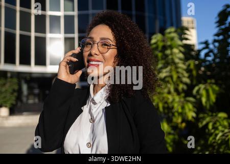 Eine selbstbewusste Geschäftsfrau mit lockigem Haar, die einen schwarzen Blazer und eine schwarze Brille trägt, lächelt, während sie draußen mit ihrem Telefon spricht, mit einem modernen Bürogebäude Stockfoto