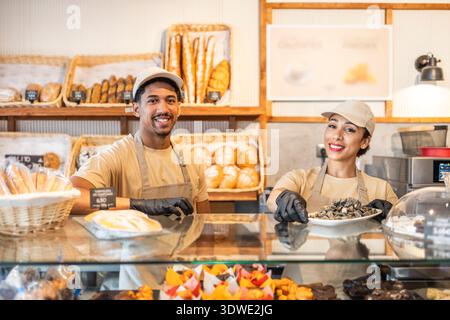 Zwei lächelnde Bäckerei-Arbeiter in Schürzen und Kappen servieren frisches Gebäck und Brot hinter einer Glasvitrine in einem gemütlichen Café. Körbe mit verschiedenen Backwaren sind vorhanden Stockfoto