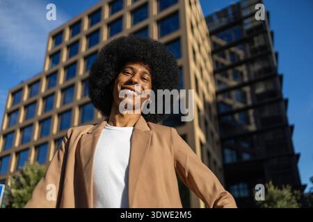 Eine Person mit braunem Blazer lächelt selbstbewusst vor der Kulisse eines modernen Bürogebäudes. Der klare blaue Himmel verstärkt die Lebendigkeit und den Erfolg Stockfoto