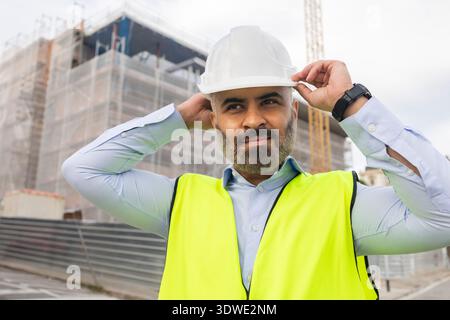 Ein Ingenieur mit weißem Schutzhelm und Neonweste passt seine Ausrüstung auf einer viel befahrenen Baustelle an. Hinter ihm ist ein Bauprojekt im Gange Stockfoto