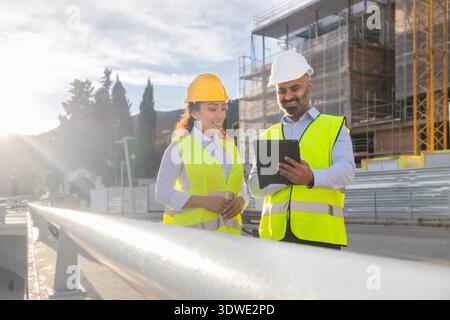 Zwei Bauingenieure in Sicherheitswesten und Helmen prüfen die Baupläne auf einem Tablet. Sie stehen in der Nähe einer städtischen Baustelle und zeigen Teamwork Stockfoto