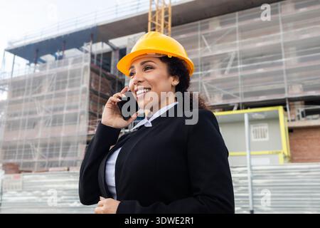 Eine Ingenieurin in formeller Kleidung und Schutzhelm spricht am Telefon und überwacht eine Baustelle. Der Hintergrund zeigt Gerüste und Baumateri Stockfoto